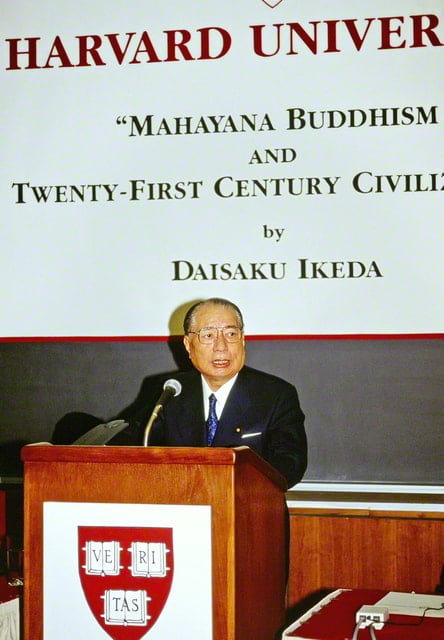 画像・講演する池田先生。背後の黒板には講演のタイトルが貼られ、演台には大学のエンブレムが大きく貼られている。