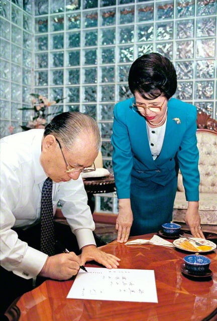 画像・スーツの上着を脱ぎ、前屈みになりながら茶色のテーブルに置かれた色紙にサインペンで書き込んでいる池田先生。その横でテーブルに手をついて見守る香峯子夫人
