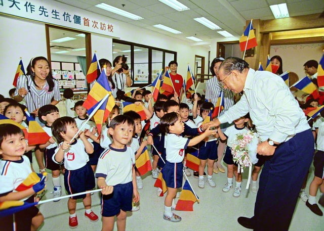 画像・三色旗をもった園児に囲まれる先生。左手に花束をもち、園児のひとりと握手しようとしている